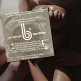 A packet of "bCondoms Extra Strength Lubricated Condoms" with a comfort fit design rests on the bare skin of two people lying close together, partially enveloped in shadows. The text on the packet includes a warning about allergic reactions and notes that using these condoms can reduce the risks of pregnancy and HIV/AIDS.